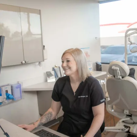Smiling female dental assistant at desk in modern dental office with equipment and X-ray monitor visible.