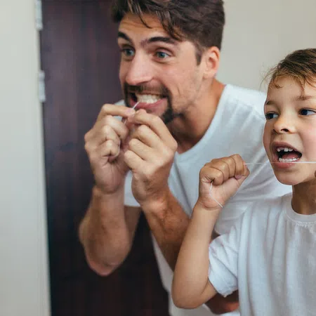 A father and son happily flossing together in a bright bathroom setting.