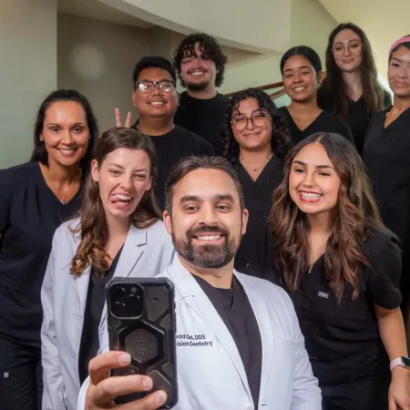 Group of smiling dental professionals taking a selfie inside a clinic with stairs and glass railing.