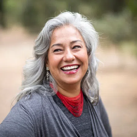Smiling woman with silver hair and red necklace in a natural setting, exuding warmth and positivity.