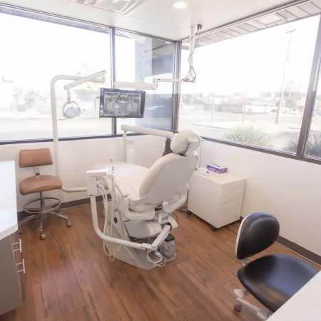 Bright dental clinic room with white dental chair, equipment, brown and black swivel chairs, and large windows.