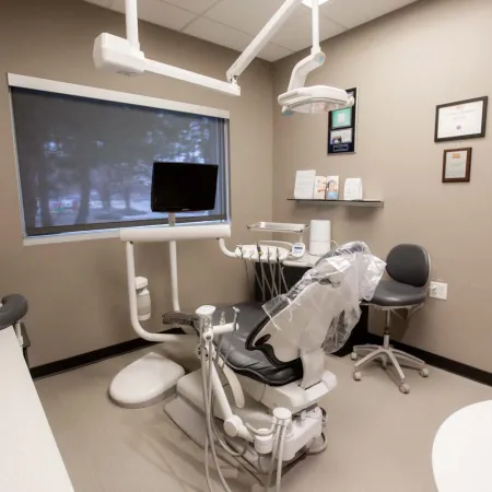 Modern dental clinic room with dental chair, equipment, computer monitor, and office chair under warm lighting.