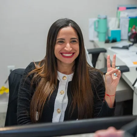 Smiling woman at office desk making a peace sign with fingers, computer and office supplies in background.