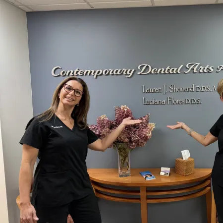 Two smiling female dentists in black scrubs pose in front of Contemporary Dental Arts P.C. sign with flowers and brochures.