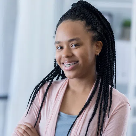 Smiling girl with braided hair and braces, standing confidently indoors with natural light.