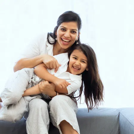 Mother and daughter smiling and hugging each other while sitting on a gray couch in bright room.