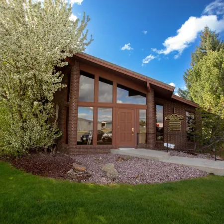 Brick office building with large windows surrounded by green trees and landscaping under a blue sky.