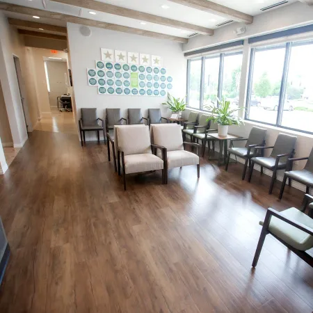 Bright waiting room with wooden floor, beige and gray chairs, plants, and large windows letting in natural light.