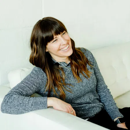 Smiling woman with long brown hair sitting relaxed on a white couch wearing a gray sweater and black pants.