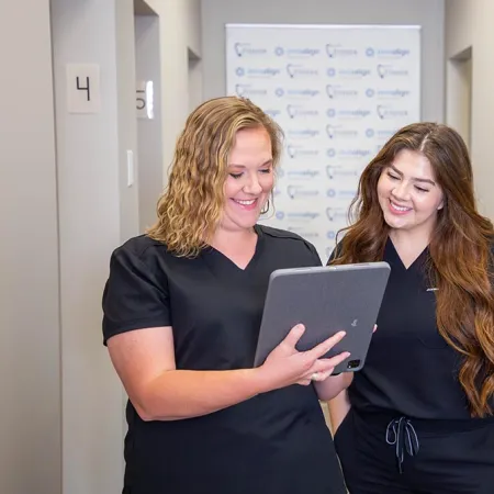 Two smiling female healthcare professionals in black scrubs reviewing a tablet in a medical office hallway.
