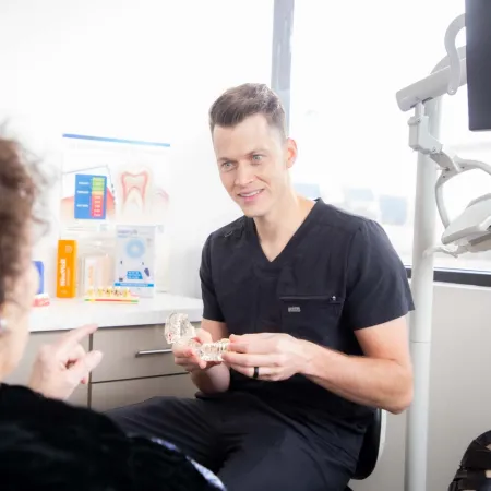 Dentist explaining dental model to patient in bright clinic with tooth chart and dental tools.