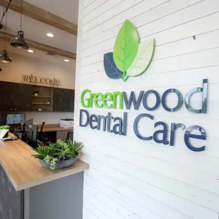 Bright dental office reception with Greenwood Dental Care sign and a wooden desk with plants and computers.