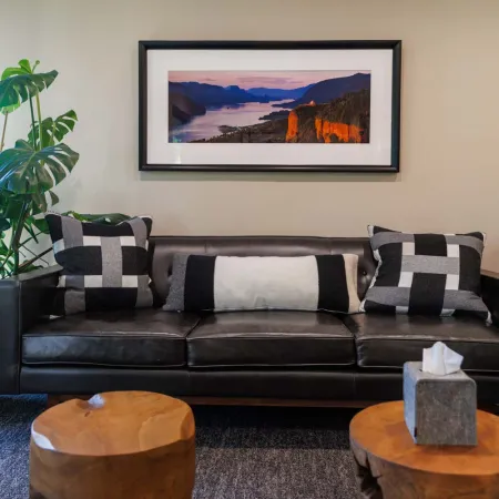 Modern living room with black leather sofa, geometric pillows, wooden stools, large plant, and framed landscape photo