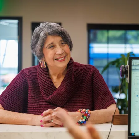 Smiling senior woman in maroon sweater engaged in conversation at a desk with calendar on computer screen.