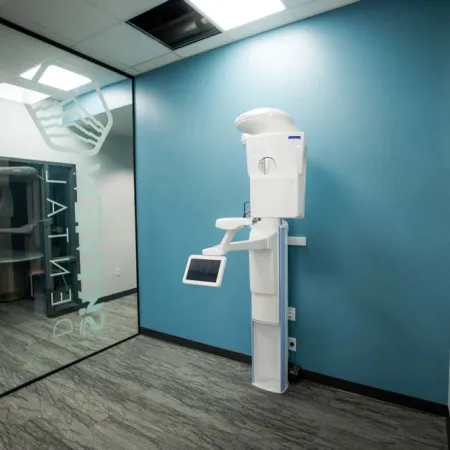 Modern dental X-ray machine mounted on blue wall in a clean, minimalist dental clinic room with glass partition.