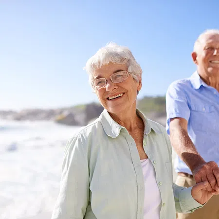 Happy elderly couple holding hands at the beach with a sunny sky and gentle waves in the background.