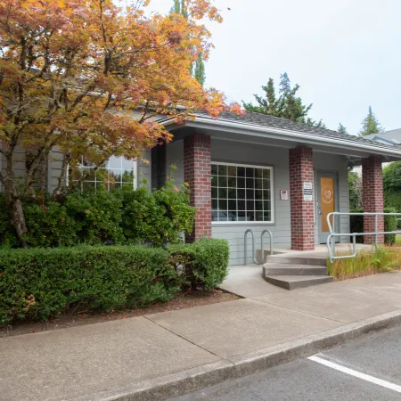 Small building entrance with brick pillars, wheelchair ramp, and autumn foliage beside sidewalk and parking lot.