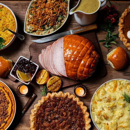Thanksgiving feast with glazed ham, pecan pie, pumpkin pie, casseroles, dinner rolls, gravy, and candles on wooden table.