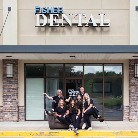 Dental team in black uniforms posing on a chair outside Fisher Dental clinic with building signage visible