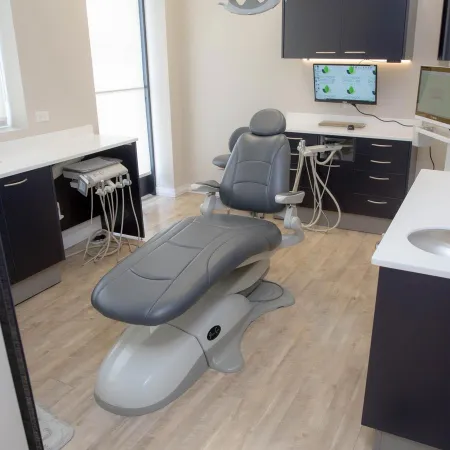 Modern dental clinic room with a gray dental chair, equipment, and monitors on light wood flooring.