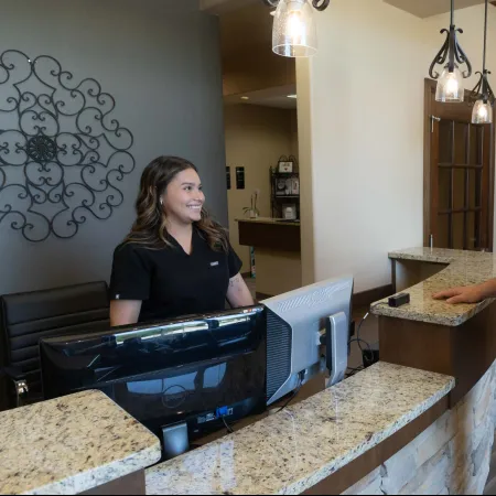 Receptionist in black scrubs smiling at a male client at a clinic's granite reception desk with computers