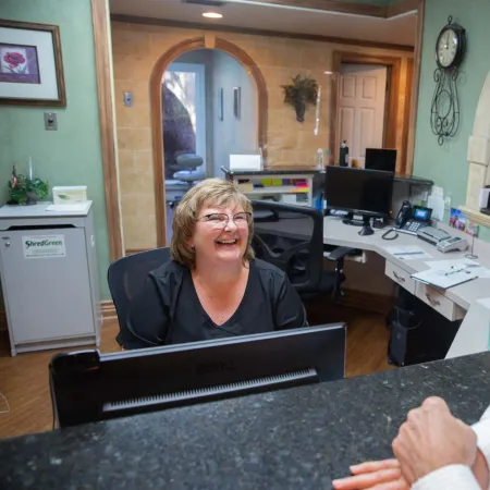 Smiling receptionist talking to a male visitor at the front desk in a warmly lit office lobby.
