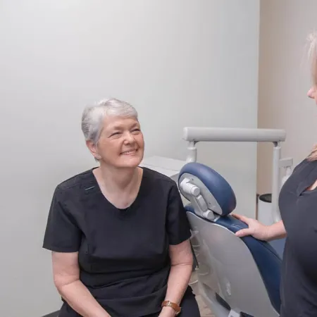 Two female dental professionals in black scrubs smiling and conversing near a dental chair in a clinic.