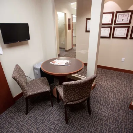 Small office room with round table, two patterned chairs, wall-mounted TV, and framed certificates on the wall.