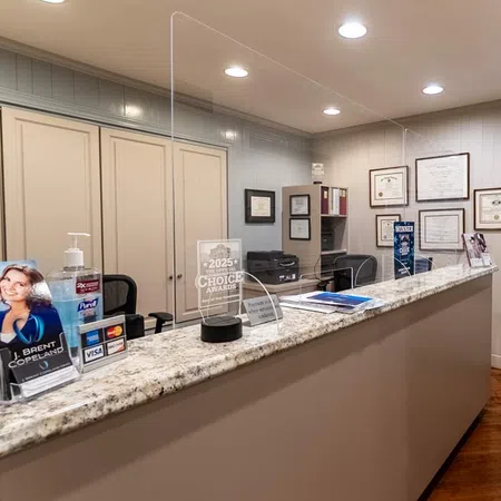 Reception desk with clear protective barrier, awards, hand sanitizer, and framed certificates on office wall.
