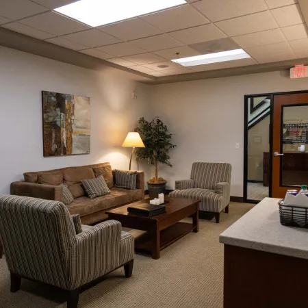 Cozy office lounge with a brown sofa, two striped chairs, wooden coffee table, lamp, and wall art.