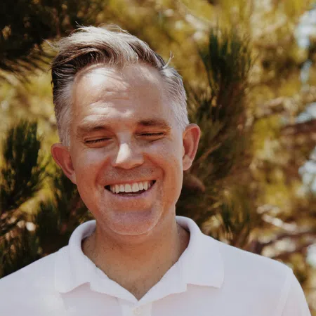 Smiling middle-aged man with gray hair wearing a white polo shirt outdoors with trees in background