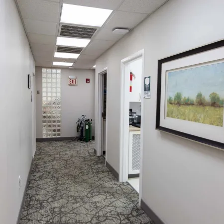 Clean office hallway with carpet, framed landscape painting, fluorescent ceiling lights, and exit sign at the end.