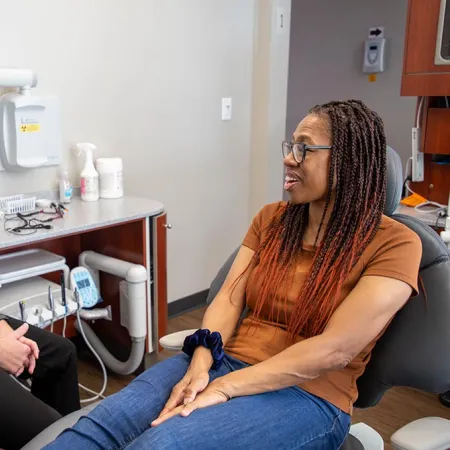 Dentist in black scrubs talking to a woman patient sitting in a dental chair in a clinic room.
