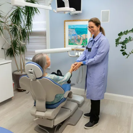 Dentist in a blue coat shaking hands with a patient seated in a dental chair in a modern clinic.