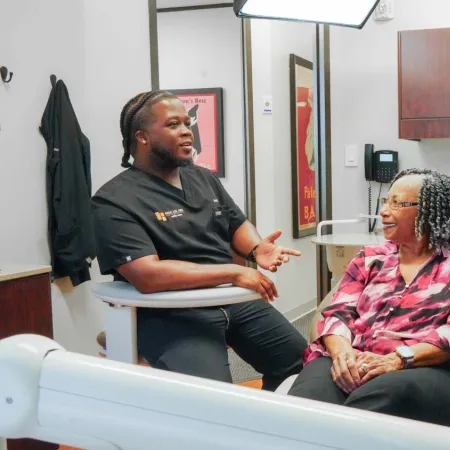 Two dentists engage with a smiling female patient in a modern dental office with wooden cabinets and a monitor.