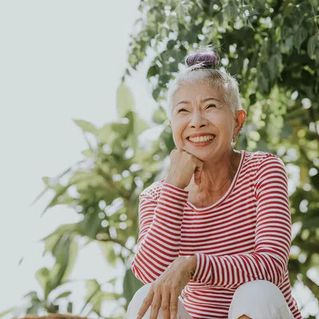 Smiling senior woman sitting outdoors in a striped shirt with greenery in the background.