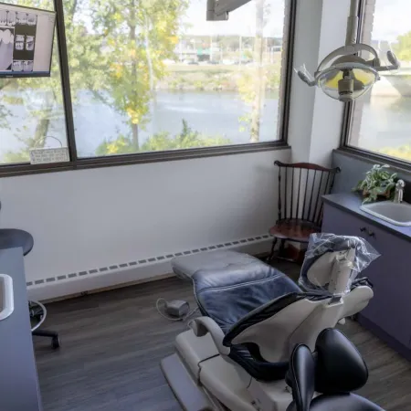 Modern dental clinic room with operatory chair, X-ray monitor, large windows overlooking a river landscape.