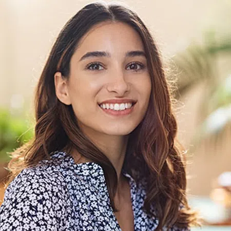 Smiling young woman with long brown hair wearing a floral blouse, sitting outdoors in soft natural light.