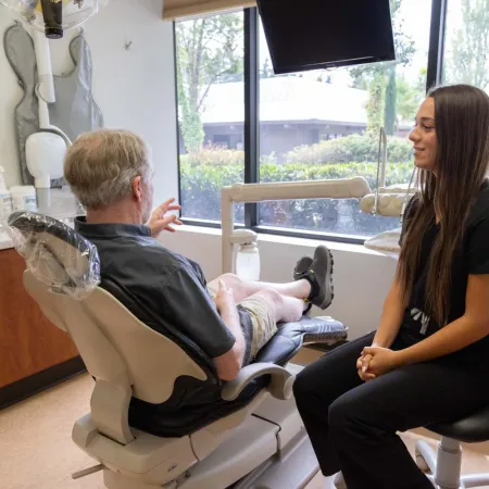 Dentist consulting an elderly male patient in a bright dental office with large windows and modern equipment.