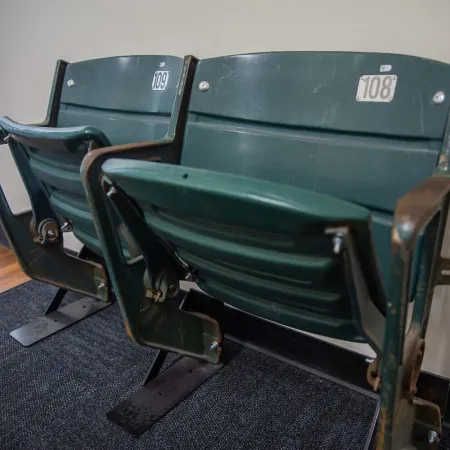 Vintage green stadium seats numbered 108 and 109 mounted indoors on carpet near window.