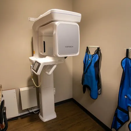 Modern dental X-ray machine in a small room with two blue lead aprons hanging on the wall.