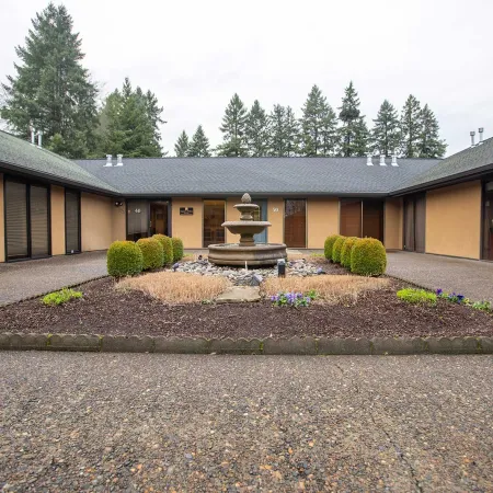 Courtyard with central stone fountain surrounded by manicured bushes and low beige buildings under an overcast sky.