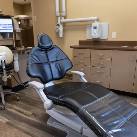 Modern dental office with black reclining dental chair, computer, stools, and beige cabinets under bright lighting.