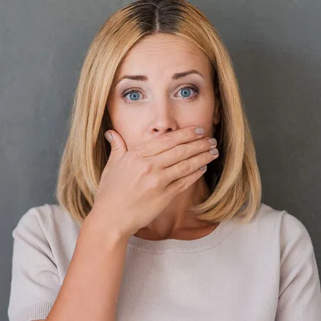 A woman with blonde hair covers her mouth, showing a surprised expression against a gray background.