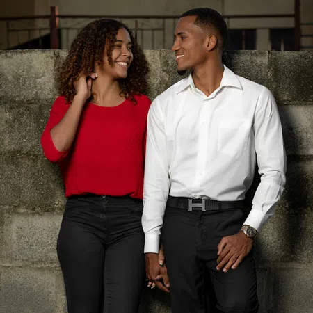 Young couple holding hands and smiling at each other while standing against a concrete wall outdoors.