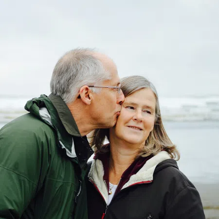 Senior man in green jacket kisses his smiling partner on the cheek at a beach with overcast sky and ocean waves.