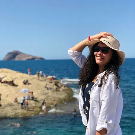 Woman in sunglasses and sunhat smiling by the rocky sea shore on a sunny day with ocean and island in background