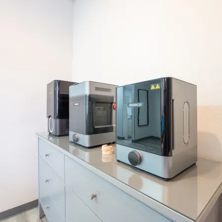 Three advanced desktop 3D printers on a gray cabinet in a bright room with white walls and tiled floor.