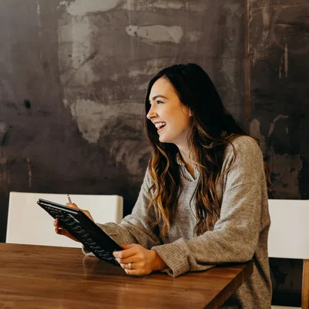 Young woman in cozy sweater smiling and holding a tablet while sitting at a wooden table in a modern room