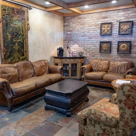 Cozy living room with brown leather sofas, floral armchair, brick wall, ornate tapestry, and warm lighting